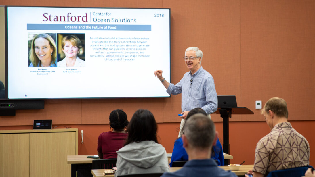 A man stands in front of an audience presenting slides. The wall behind him is orange.