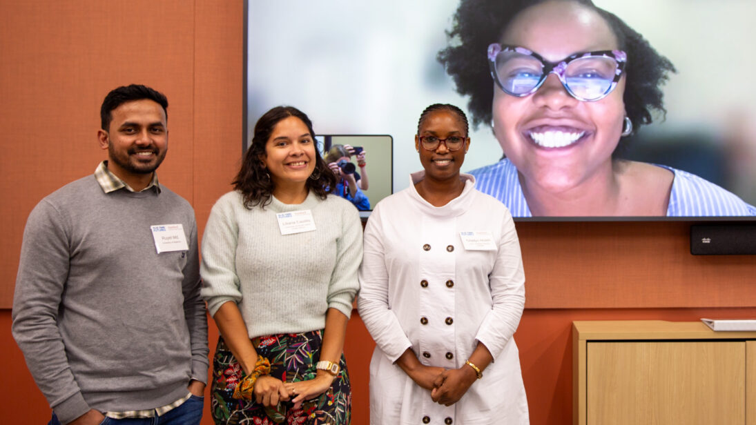a group of people pose in a conference room in front of a screen, where another person is posing remotely