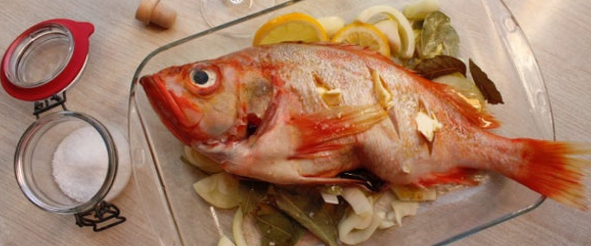 A glass dish on a table with a whole baked fish over vegetable.s next to a jar of salt