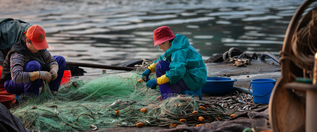 Two women sit on the harborside untangling fishing nets.