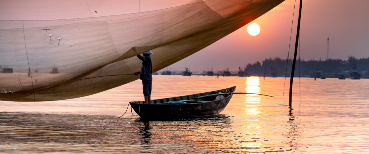 Artisan fisher in a small wooden boat tends to nets in front of a beautiful sunset.